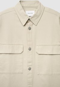 Khakiowa koszula z długim rękawem, zapinana na guziki z przodu, z dwoma kieszeniami na piersi, kołnierzykiem w szpic i srebrzystymi guzikami. Wykonana z bawełnianej tkaniny o fakturze.