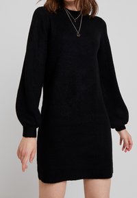 Robe pull noire en maille avec un col rond, de longues manches ballon et une silhouette ajustée, dotée de poignet et d'ourlet côtelés. Texture douce.