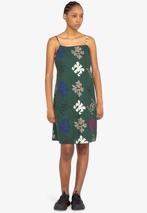 Vestido verde sin mangas con patrones florales en púrpura, blanco y beige. Cuenta con tirantes finos y un corte relajado. Se lleva con calzado negro.