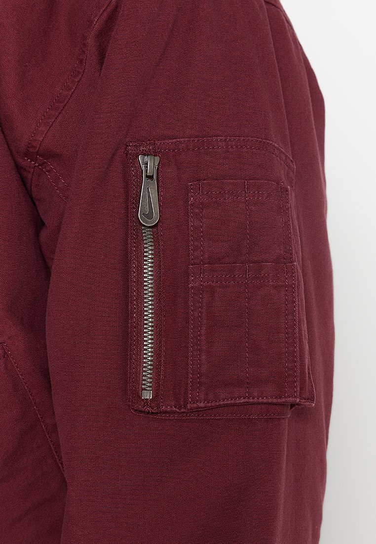 Manica di giacca in tessuto bordeaux con una tasca con zip e un tirante argentato, e una tasca applicata, entrambe con cuciture rinforzate.