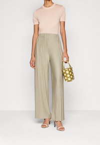 Svetle růžový úpletový top s krátkým rukávem je kombinován s plisovanými, širokými béžovými kalhotami; modelka drží malou, texturovanou zelenou kabelku ve tvaru kbelíku.