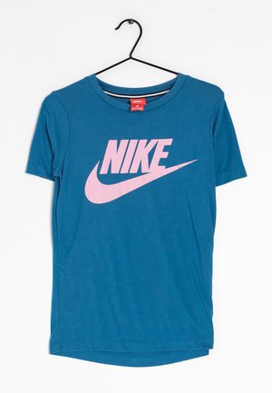 Modré tričko s krátkým rukávem s velkým růžovým logem Nike a znakem swoosh, visící na černém ramínku na bílém pozadí.