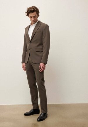 Giovane uomo con un abito marrone, camicia bianca e mocassini neri, in piedi contro uno sfondo beige semplice, guarda verso il basso.