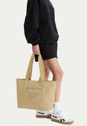 Osoba ubrana w czarne szorty i bluzę, trzymająca beżową torbę typu tote marki Reebok z przednią kieszenią na zamek, białe skarpetki i białe buty sportowe Reebok.