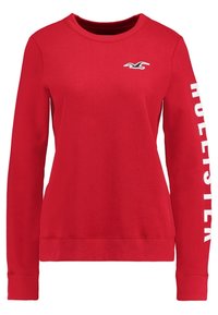 Felpa rossa con maniche lunghe, scollatura rotonda, polsini e orlo a coste. Presenta la scritta bianca "HOLLISTER" lungo una manica e un logo sul petto.