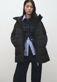 Czarna pikowana kurtka z kapturem, z zamkiem błyskawicznym i przednimi kieszeniami. Nosi się ją na niebieskiej koszuli w paski i dżinsowej kurtce.