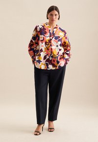 Blusa a stampa floreale colorata con maniche lunghe, colletto rialzato e tasche, abbinata a pantaloni neri su misura e sandali neri con tacco.