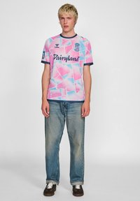 Kurzärmliges, pink- und blau gemustertes Sporttrikot mit marineblauen Akzenten, runder Ausschnitt und bedrucktem Text. Getragen mit hellblauen Jeans.