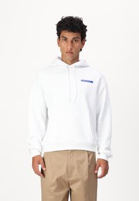 Felpa bianca con cappuccio, caratterizzata da un logo blu sul petto, tasca frontale e polsini a costine. Abbinata a pantaloni khaki.