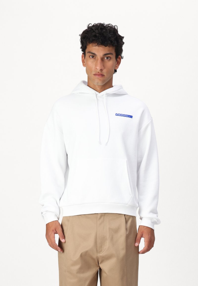 Felpa bianca con cappuccio, caratterizzata da un logo blu sul petto, tasca frontale e polsini a costine. Abbinata a pantaloni khaki.