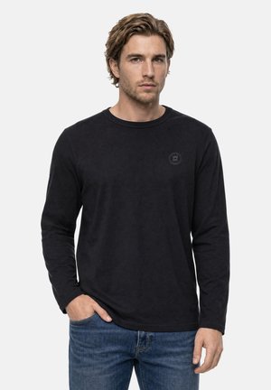 Homme aux cheveux châtain clair portant une chemise noire à manches longues et un jean bleu, debout avec une main dans la poche devant un fond blanc.