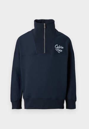 Pull-over en coton bleu marine avec un col montant, fermeture à moitié éclair, et logo Calvin Klein blanc sur la poitrine. Comprend des poignets et un ourlet côtelés.