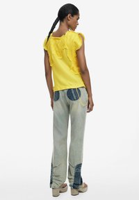 Gelbes Oberteil mit strukturiertem Blumendetail, kombiniert mit hellblauen Jeans mit dunkleren Flecken und ausgestellten Beinen, ergänzt durch klobige Absätze.