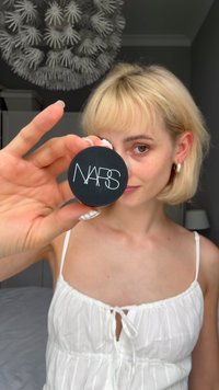 Compact NARS avec un conteneur noir rond arborant un logo blanc. Fini lisse, texture glossy, tenu par une personne aux ongles de couleur claire.