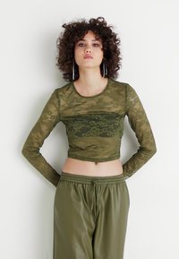 Olivgrön långärmad spetscroptop med blommönster, i kombination med matchande högmidjade byxor. Genomskinlig textur, åtsittande design.