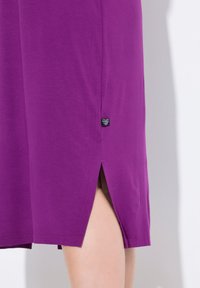 Vestido morado con una abertura lateral, hecho de una tela suave. Incluye una pequeña etiqueta cerca del dobladillo, que exhibe un diseño simple.