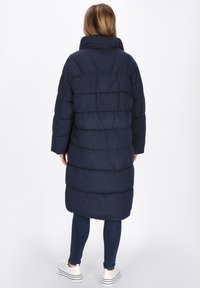 Persona che indossa un lungo cappotto imbottito blu navy, jeans scuri e sneaker bianche con plateau, in piedi di spalle rispetto alla telecamera su uno sfondo bianco uniforme.