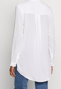 Chemise blanche à manches longues avec poignets boutonnés et pli dans le dos, portée non rentrée sur un jean bleu.