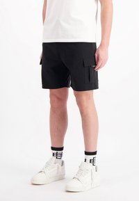Zwarte cargoshorts met zijzakken, gemaakt van lichte stof. Gecombineerd met witte sneakers en gestreepte zwart-witte sokken.