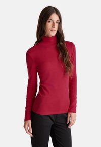 Frau mit langen braunen Haaren, die einen roten Rollkragenpullover und schwarze Hosen trägt, steht vor einem weißen Hintergrund.