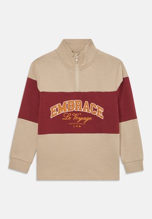 Beige og burgunder halv-zip hettegenser med lange ermer. Har broderte teksten "EMBRACE Le Voyage" i gult. Laget av mykt bomullsmateriale.