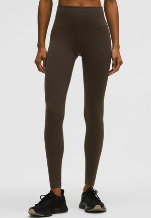 Frau trägt hoch taillierte dunkelbraune Leggings und schwarze Sportschuhe, steht vor einem schlichten hellen Hintergrund.