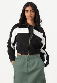Čierna crop bunda na zips s bielym diagonálnym pruhom a zlatým logom; skombinovaná s zelenými cargo nohavicami na šnurovanie. Hladká textúra látky.