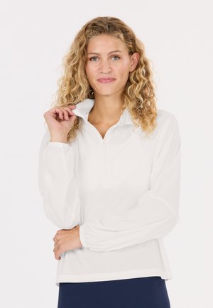 Frau mit lockigen blonden Haaren, die einen weißen Langarmpullover trägt, hält mit einer Hand den Kragen und hat den anderen Arm verschränkt.