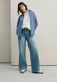 Camicia a righe blu e bianche con bottoni sopra una maglietta bianca, abbinata a pantaloni di jeans a gamba larga di un blu chiaro e sandali bianchi con suole nere.