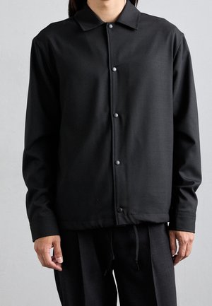 Persona que lleva una camisa negra de manga larga abotonada con cuello y pantalones negros a juego, de pie contra un fondo claro y liso.