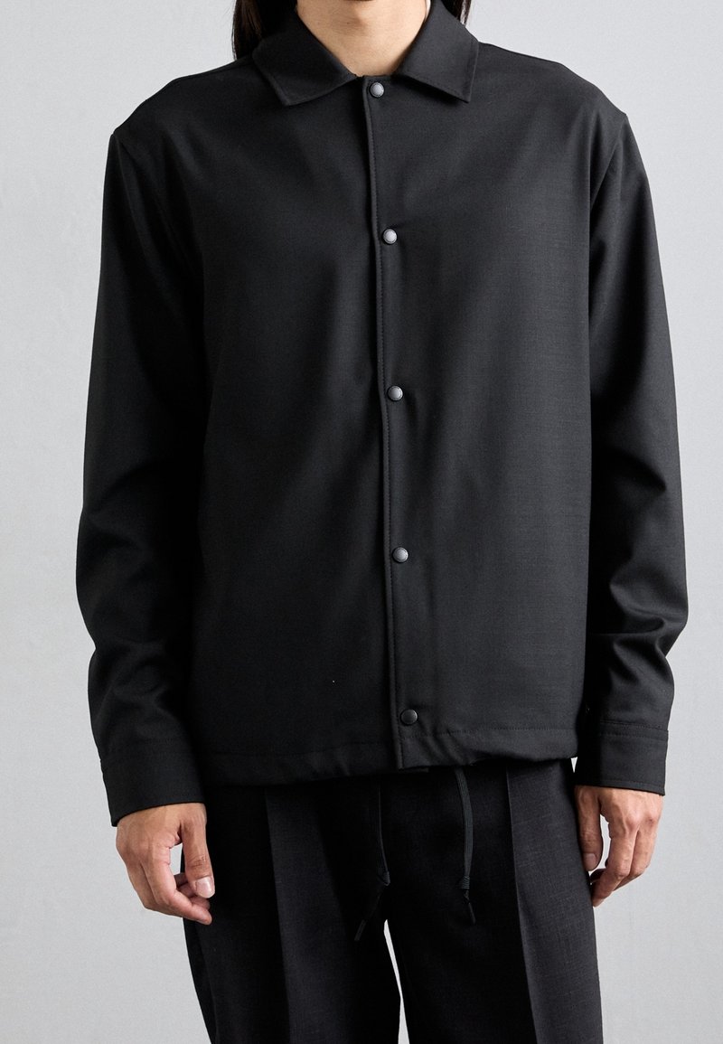 Personne portant une chemise noire à manches longues boutonnée avec col et un pantalon noir assorti, se tenant devant un fond clair uni.