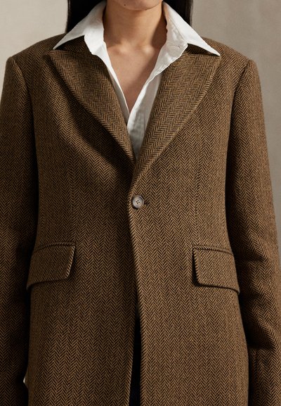 Kobieta nosząca brązową marynarkę w jodełkę na białej koszuli z kołnierzykiem, marynarka jest zapięta na guziki, widoczne są przednie kieszenie z patkami.