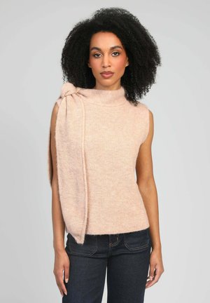 Femme aux cheveux bouclés portant un haut beige sans manches en tricot avec un grand nœud sur l'épaule et un jean foncé, debout devant un fond gris uni.