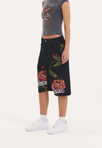 Shorts de mezclilla negra con estampados florales en rojo y verde, con un corte holgado y bolsillos laterales. Combinados con zapatillas blancas.