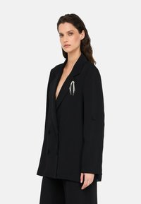 Blazer doppiopetto nero con texture liscia, caratterizzato da un rever a incastro e una spilletta argentata sul lato sinistro.