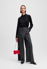 Camicia nera con colletto, abbinata a pantaloni larghi a righe grigie. Accessoriata con una borsa trapuntata rossa. Scarpe nere a punta.