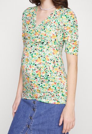 Těhotenská halenka s květinovým potiskem v zelené, žluté a růžové barvě, se zavinovacím designem a krátkými rukávy, v kombinaci s džínovou sukní.