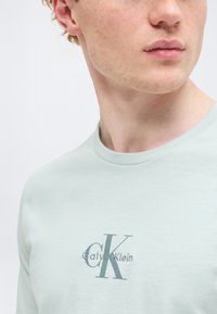 Νέος άνδρας που φοράει ανοιχτοπράσινο t-shirt Calvin Klein, κομμένο στο πηγούνι και στο στήθος, με το λογότυπο στο κέντρο του πουκάμισου.