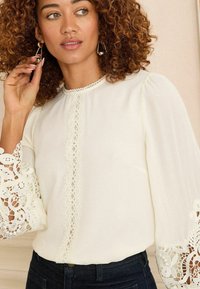Cream blouse with intricate lace trim on sleeves and front, round neckline, and long puff sleeves; paired with dark denim jeans.