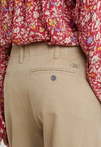 Beiga byxor med en slät textur, som har en bakficka med en knapp och en logotyp. Sömmen är snyggt avslutad.