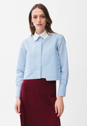 Frau trägt ein hellblaues Langarmshirt mit verziertem weißem Kragen und einem hochtaillierten dunkelroten Rock, steht vor weißem Hintergrund.