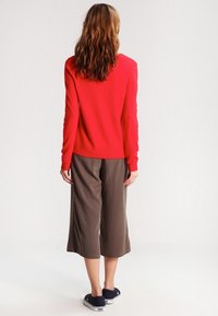Czerwony wełniany sweter z długimi rękawami, w zestawieniu z luźnymi brązowymi kulotami; model w ciemnoniebieskich sneakersach; widok z tyłu.