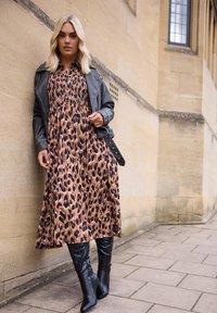 Maxi sukienka w brązowy wzór panterki, z marszczonym stanikiem, połączona z czarną skórzaną kurtką i wysokimi czarnymi botkami na obcasie do kolan.