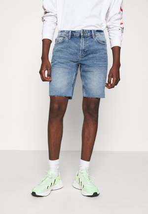 Man die lichtblauwe spijkershorts, een wit shirt met lange mouwen, witte sokken en lichtgroene sneakers draagt, staand tegen een effen achtergrond.