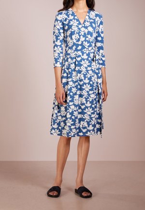 Vestido cruzado azul con estampado floral blanco, mangas tres cuartos, escote en V y falda drapeada. Combinado con sandalias planas negras.