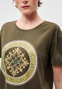 Ελαιοπράσινο βαμβακερό μπλουζάκι με κυκλική, χρυσή και πράσινη διακοσμητική εκτύπωση που περιλαμβάνει στιλιζαρισμένα μοτίβα και κείμενο σε αντίθετη γραμματοσειρά.