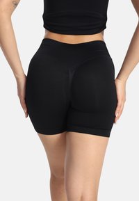 Mujer con shorts negros ajustados de talle alto y top corto, vista desde atrás sobre un fondo blanco liso.