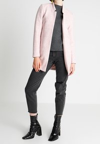 Heller rosa, strukturierten Außenmantel mit offener Vorderseite, getragen über einem dunkelgrauen Oberteil und schwarzen Jeans, kombiniert mit glänzenden schwarzen Stiefeletten.