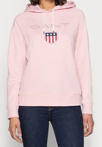 Różowa bluza z kapturem, wykonana z miękkiego materiału, z przednim logo w formie niebieskiego, czerwonego i białego herbu. Posiada sznurki w kapturze oraz ściągacze na rękawach.