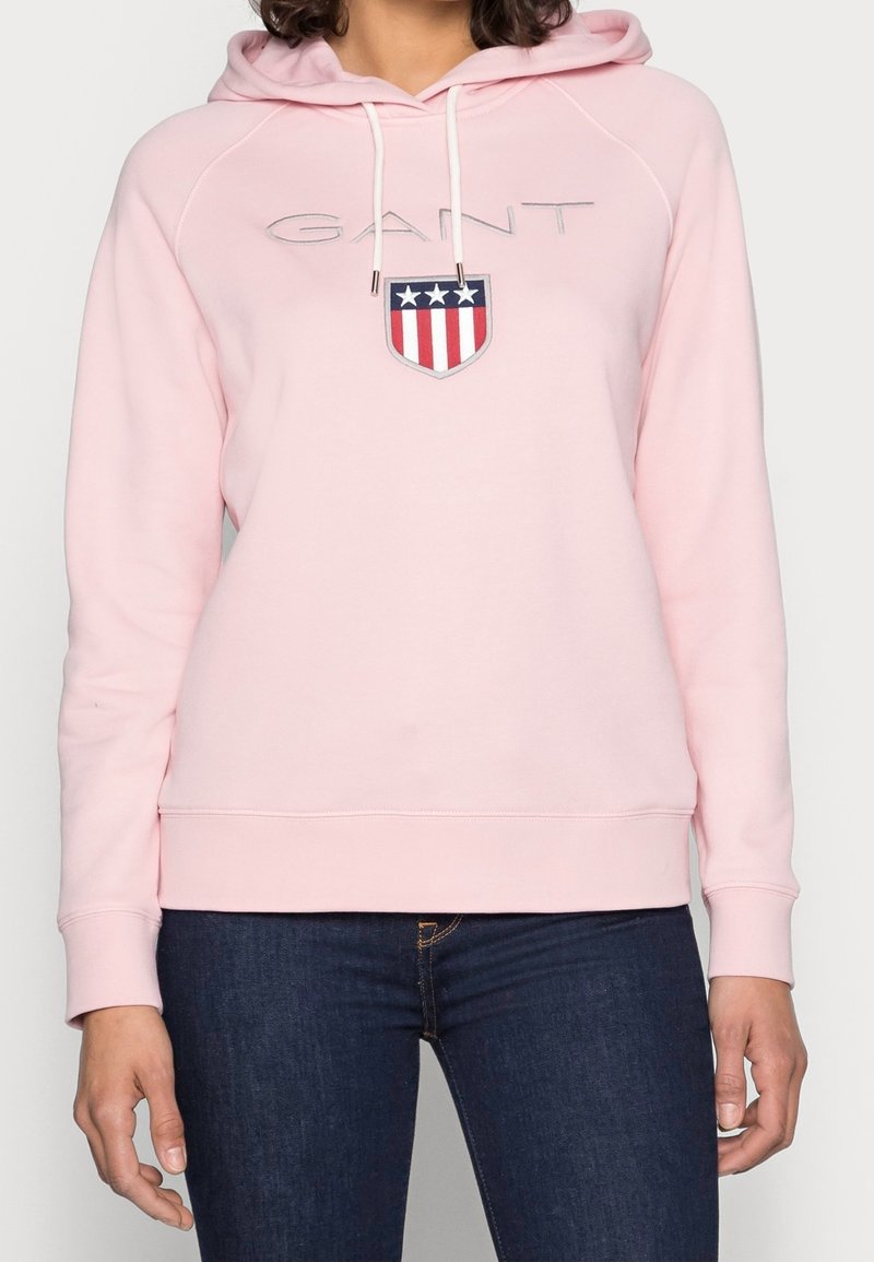Różowa bluza z kapturem, wykonana z miękkiego materiału, z przednim logo w formie niebieskiego, czerwonego i białego herbu. Posiada sznurki w kapturze oraz ściągacze na rękawach.